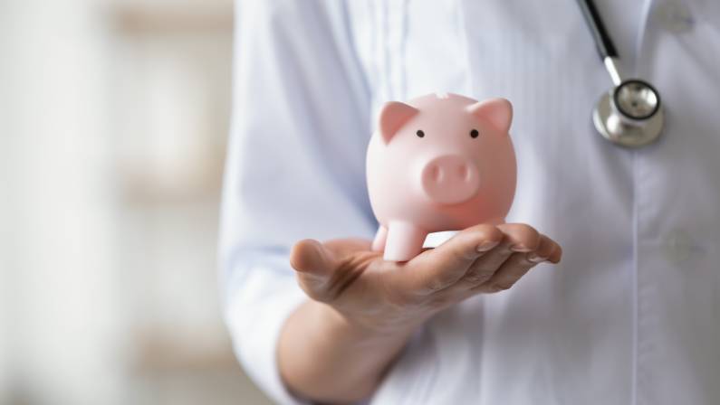 Woman doctor wearing white coat and stethoscope holding piggy bank box in hands.
