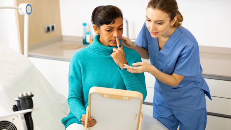 Hispanic woman holding mirror and pointing finger on her nose during cosmetologist's examination in clinic.