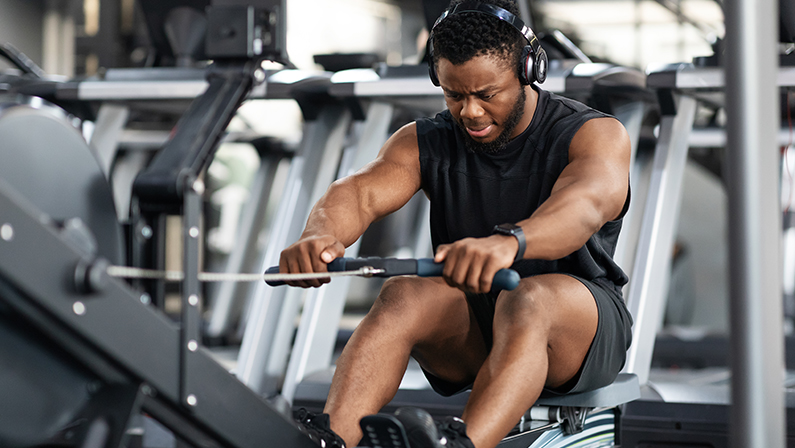 Motivated young black sportsman with wireless headset having workout on rowing machine.