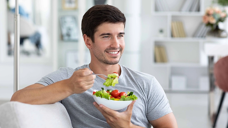 Happy man sitting on sofa at home while eating green vegetable salad. Healthy eating, restaurant dish delivery.