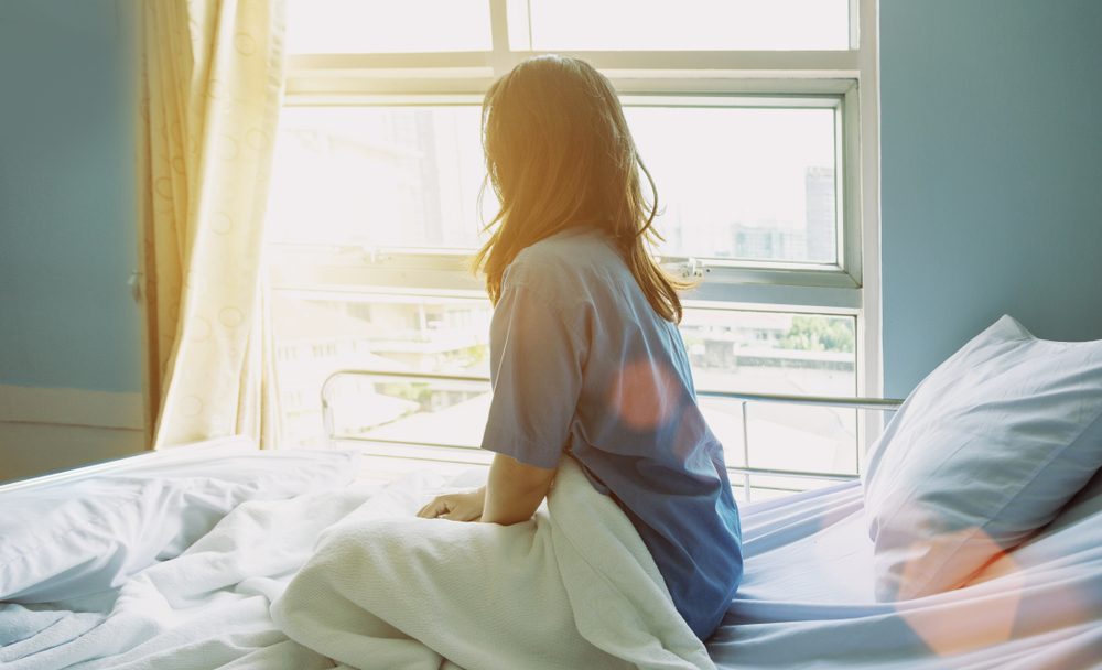 A woman in a recovery room sitting on a hospital bed, gazing out the window during her mommy makeover recovery.
