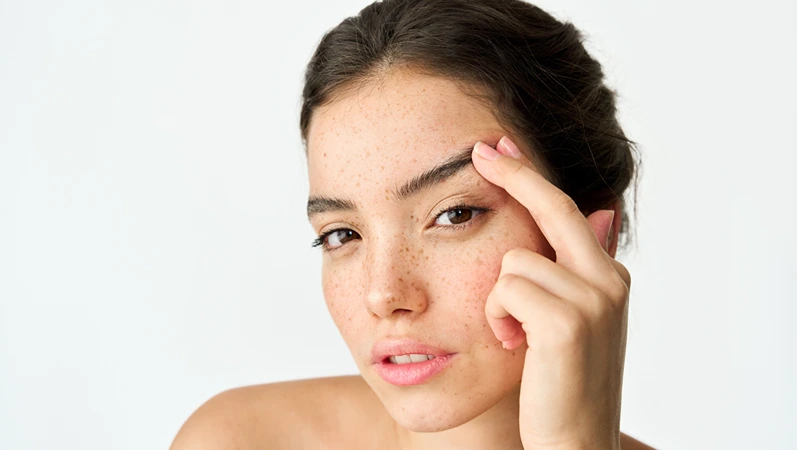 Young adult Latin woman, cool brunette gen z girl with freckles on face looking at camera lifting eyebrow isolated on white background. Beauty aesthetic portrait. Skincare, eye surgery and cosmetic ad