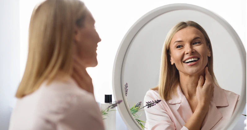Attractive Mature Lady Touching Her Smooth Skin On Neck While Looking In Mirror At Home, Beautiful Middle Aged Woman Smiling To Her Reflection, Enjoying Beauty Routine And Anti-Aging Skincare
